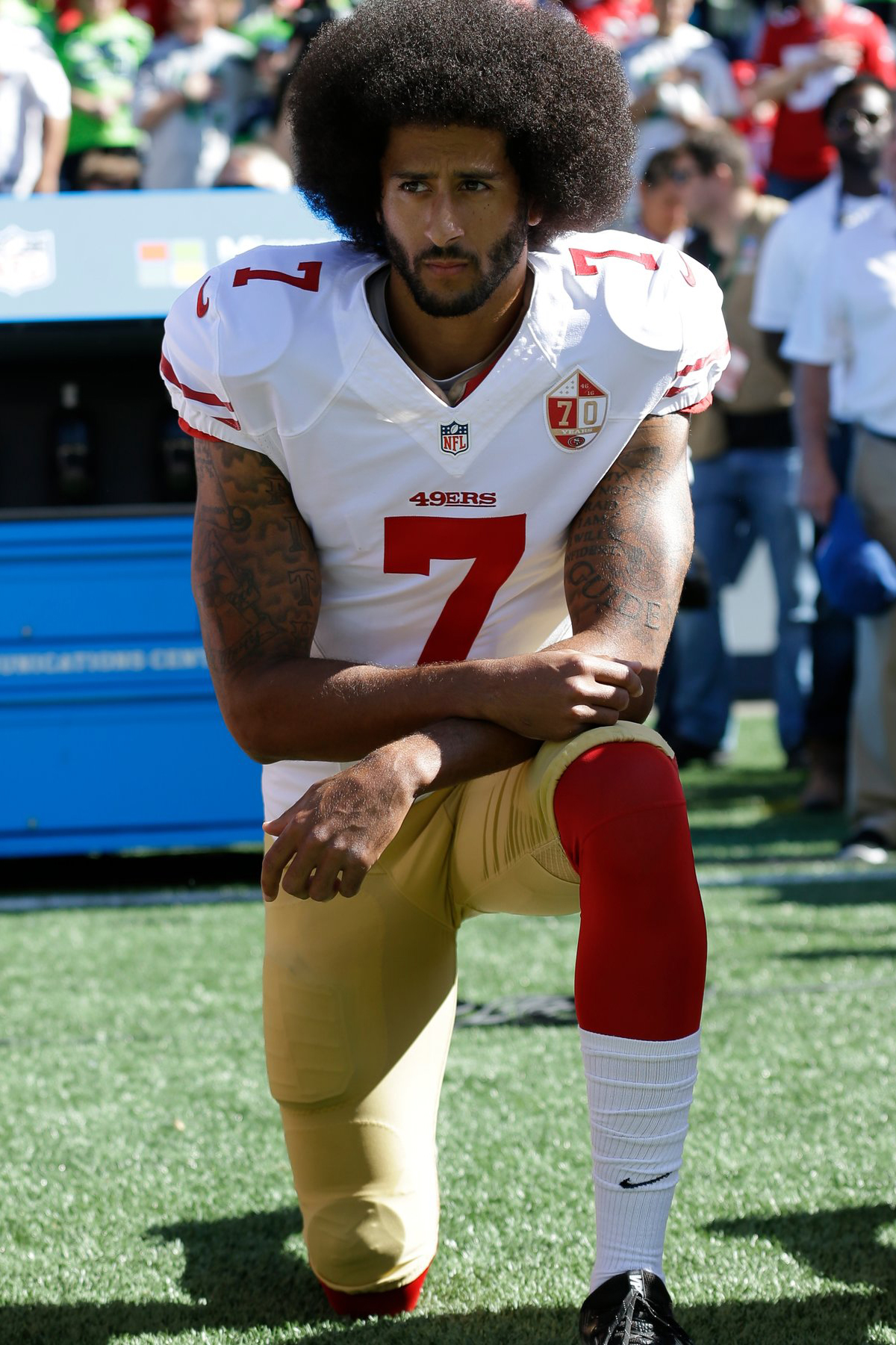 In this color photograph, Colin Kaepernick kneels on the sidelines of a football field.