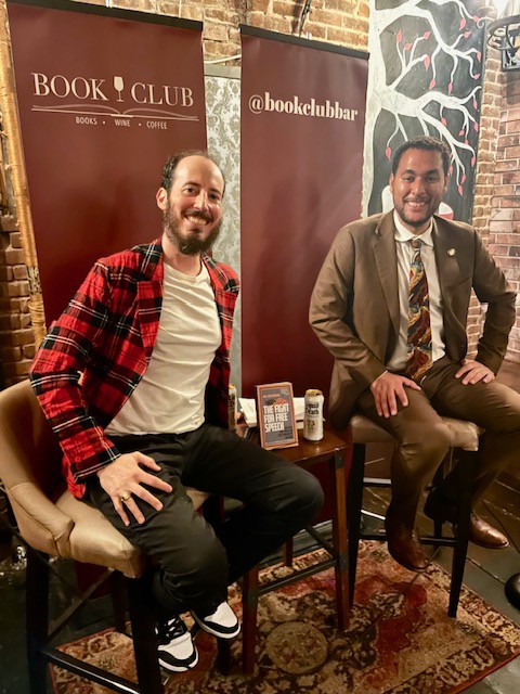 Author Ian Rosenberg poses with his book in front of a Book Club Bar banner. Beside him sits his conversation partner.