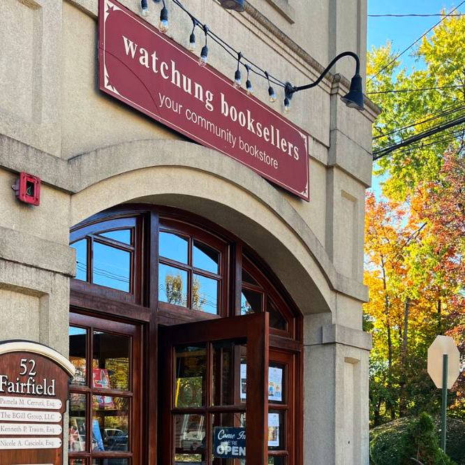 There is a beautiful stone building in front of autumn trees. Above the arched doorframe, a red sign reads Watchung Booksellers: Your Local Bookstore